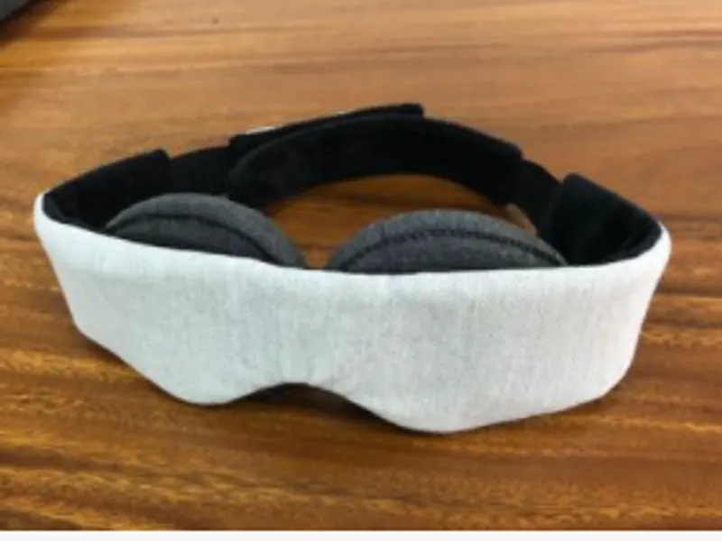 A contoured sleep mask in light grey fabric with dark eye cups and an adjustable strap, on a wooden table